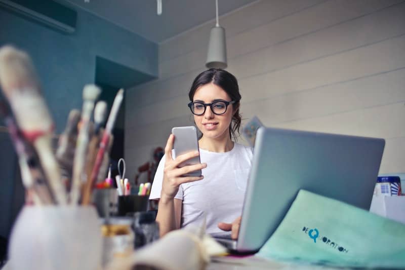 Une femme au bureau est assise derrière un ordinateur portable et tient un téléphone dans sa main