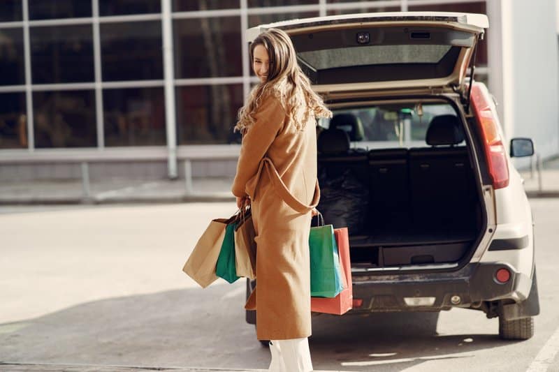 Une femme aux cheveux bruns se tient à côté d'une voiture avec des sacs à la main