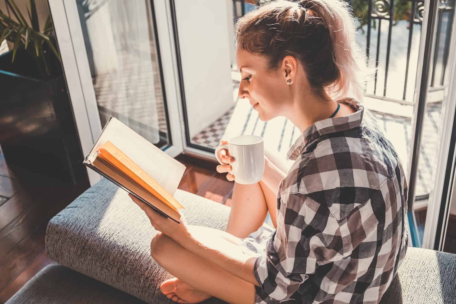 une femme aux cheveux bruns est assise près de la fenêtre en lisant un livre