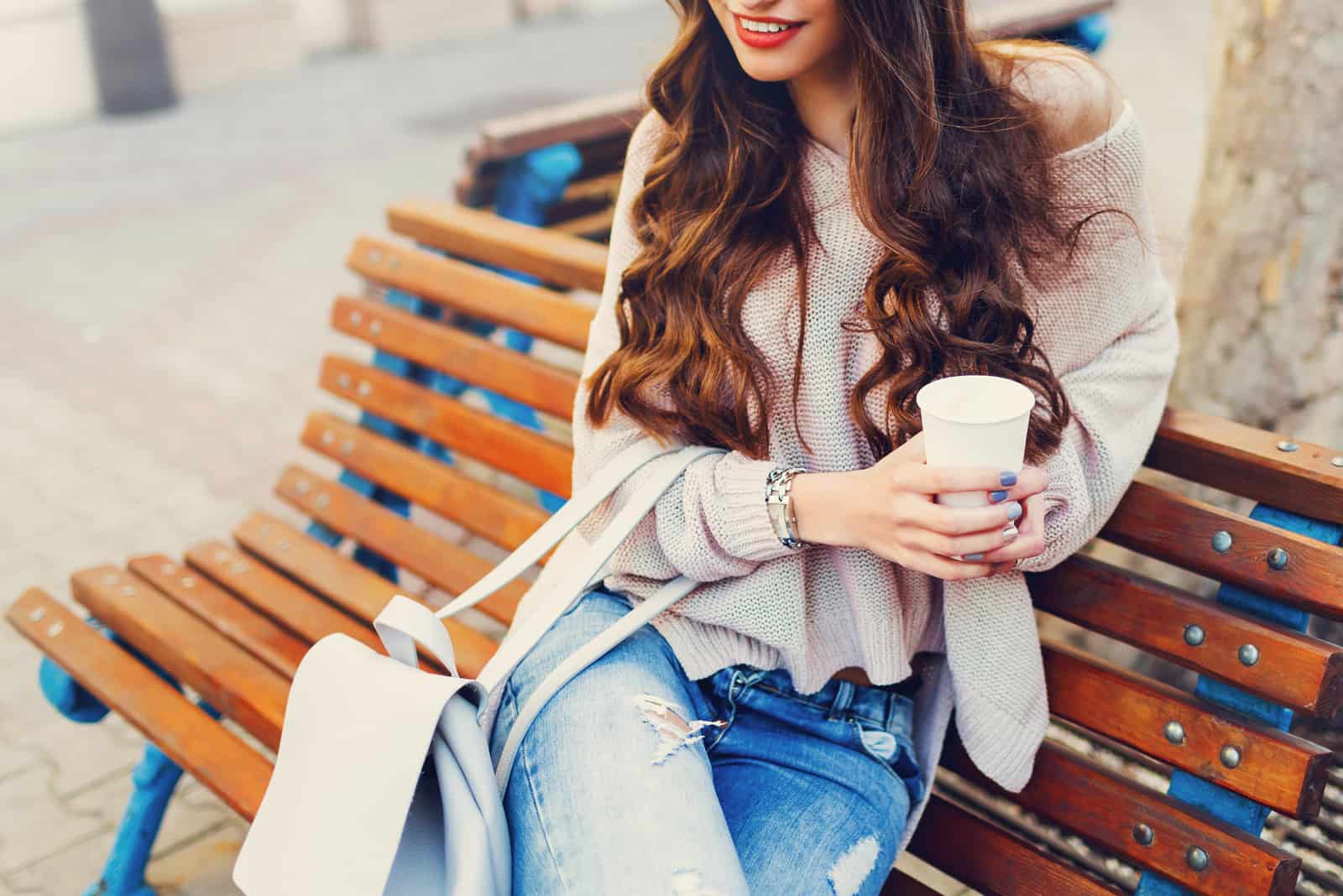 une femme aux longs cheveux bruns est assise sur un banc et tient un café à la main