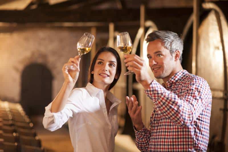 Couple dégustant un verre de vin blanc dans une cave traditionnelle entourée de fûts en bois.