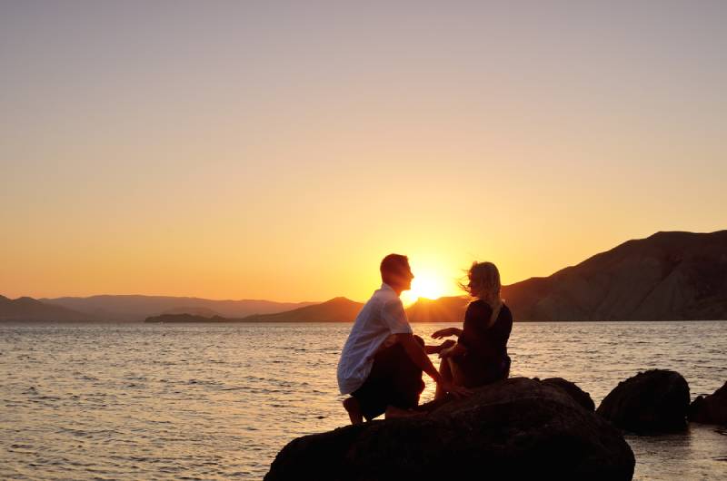 Jeune couple discutant sur un rocher au bord de la mer au soleil couchant