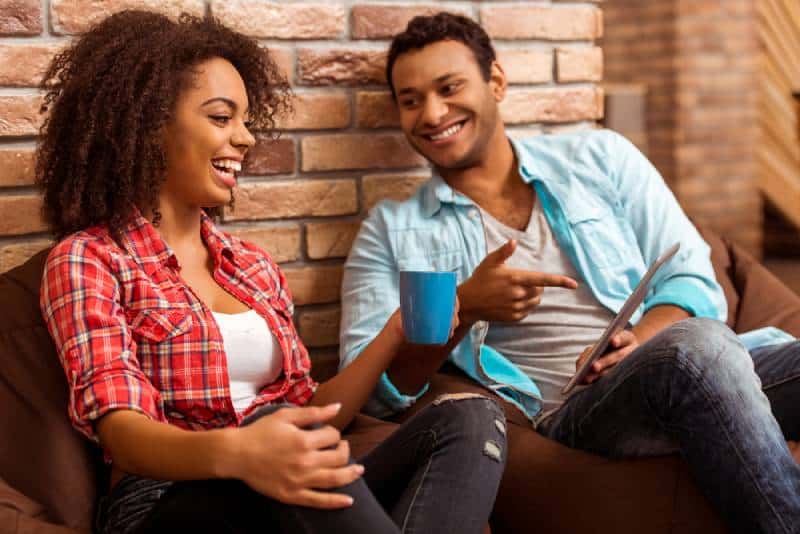 Joli couple afro-américain utilisant une tablette, tenant une tasse et riant