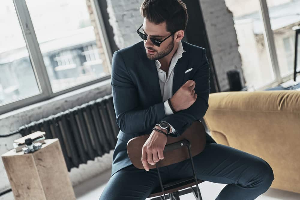 Un beau jeune homme en costume et lunettes regarde au loin alors qu'il est assis sur le tabouret