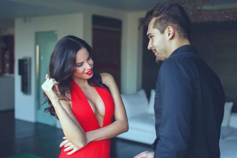 Femme en robe rouge et lys flirtant avec un jeune homme à l'intérieur, jouant avec ses cheveux