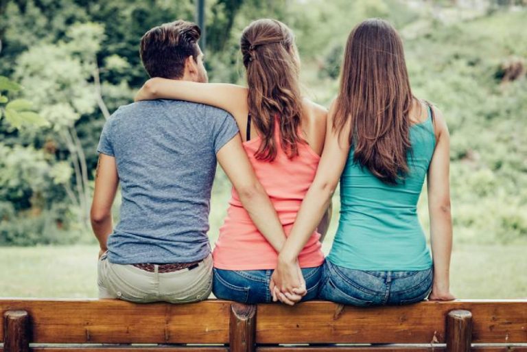 Triangle d'amour, une fille embrasse un mec et il tient la main d'une autre fille, ils sont assis ensemble sur un banc
