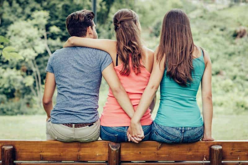 Triangle d'amour, une fille embrasse un mec et il tient la main d'une autre fille, ils sont assis ensemble sur un banc