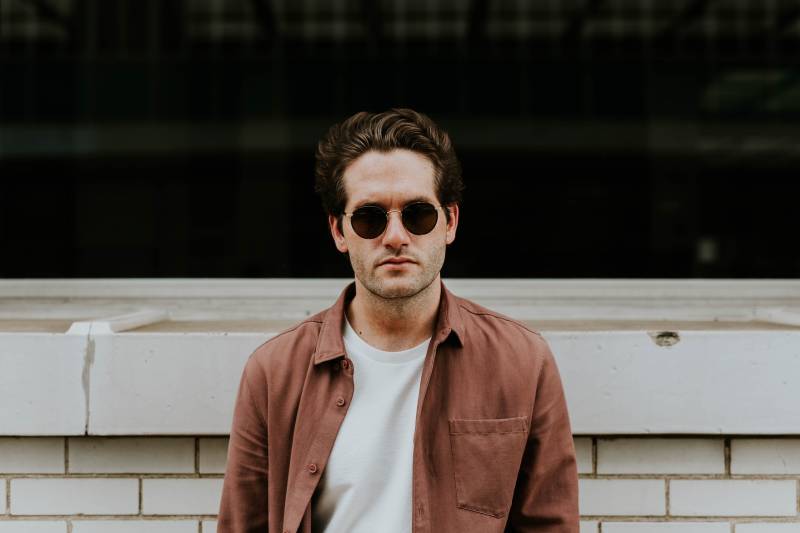Un homme avec des lunettes de soleil et une veste marron se tient à l'extérieur