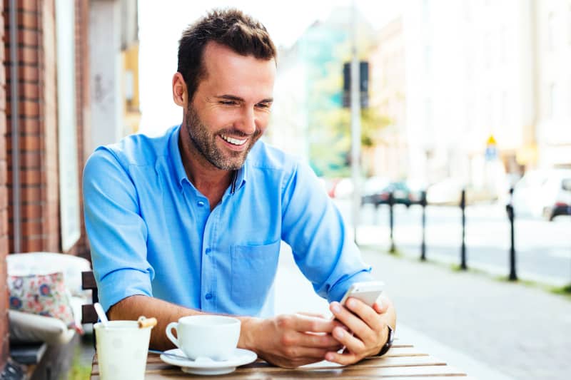 Un homme souriant en chemise bleue regarde son téléphone dans un café de rue