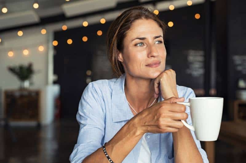 Une femme mûre et réfléchie, assise à la cafétéria, tient une tasse de café tout en détournant le regard
