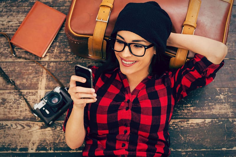 femme souriante portant une chemise à carreaux, s'appuyant sur un plancher en bois et tapant sur son téléphone