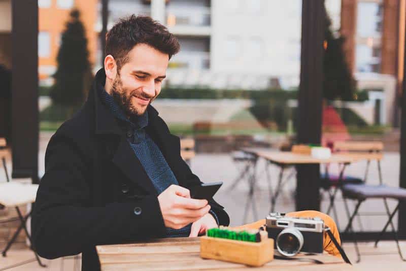 homme souriant portant un manteau noir et tapant sur son téléphone au café de la rue