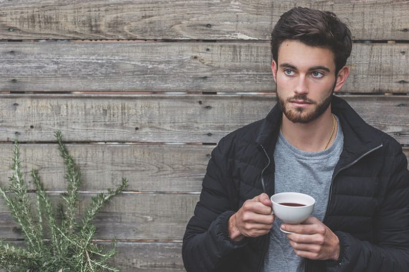 homme tenant une tasse de café en s'appuyant sur un mur de bois