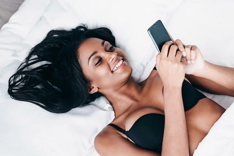 jeune femme souriante allongée sur le lit et tapant sur son smartphone