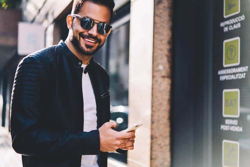 jeune homme souriant portant des lunettes de soleil et tapant sur son téléphone à l'extérieur