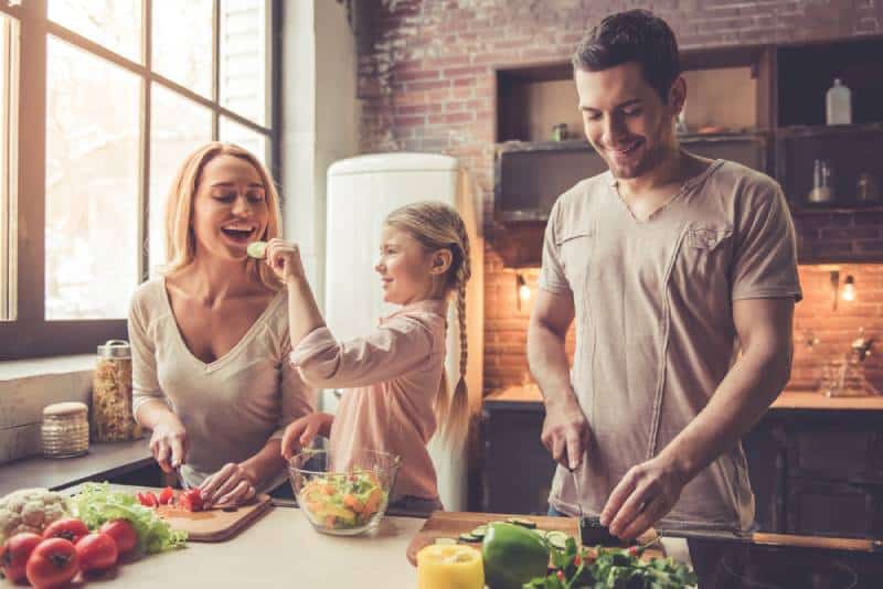Une jolie petite fille et ses beaux-parents coupent les légumes et sourient en cuisinant à la maison