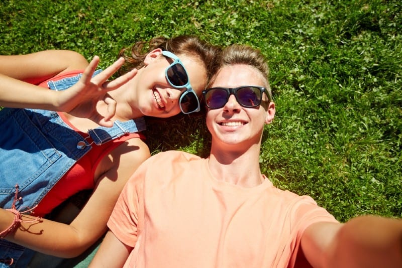 Un couple d'adolescents heureux dans des lunettes de soleil allongé sur l'herbe et prenant selfie à l'été