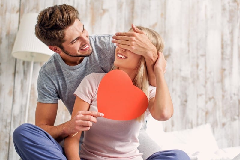 Heureux jeune homme couvrant les yeux des femmes avec surprise et lui donnant une carte coeur
