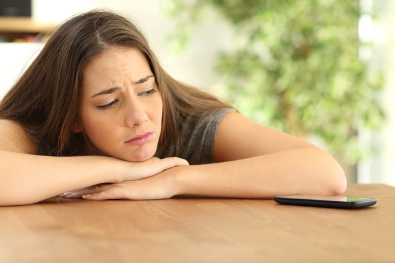 une femme triste avec sa tête sur une table en regardant un téléphone devant elle