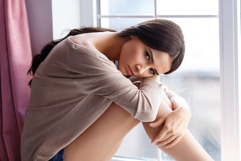 Jolie jeune fille de mauvaise humeur assise sur le rebord de la fenêtre et s'appuyant sur ses jambes tout en réfléchissant