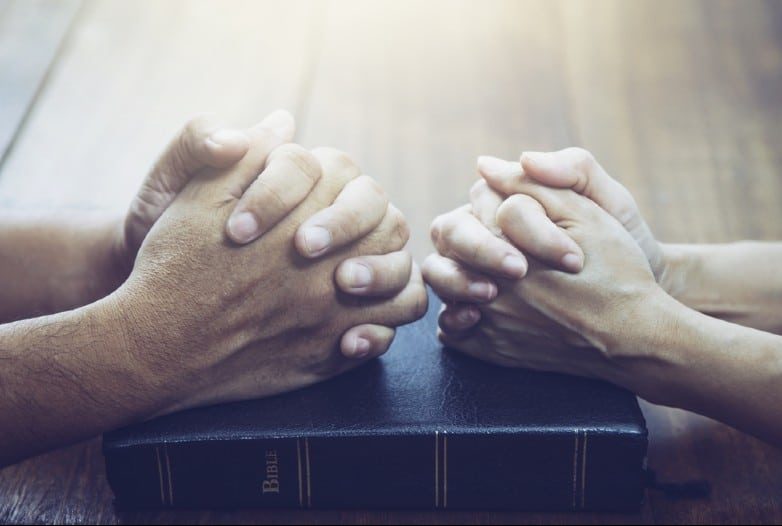 Manos de hombre y mujer rezando juntos sobre la sagrada biblia en la mesa de madera