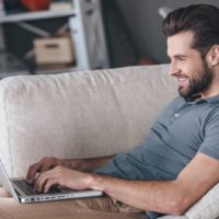 un homme souriant avec une barbe se gratte sur le canapé et correspond via un ordinateur portable