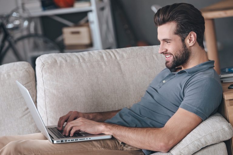 un homme souriant avec une barbe se gratte sur le canapé et correspond via un ordinateur portable