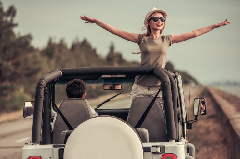 Une fille avec des lunettes de soleil se tient avec les bras tendus dans une jeep sans toit pendant la conduite