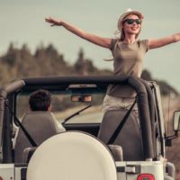 Une fille avec des lunettes de soleil se tient avec les bras tendus dans une jeep sans toit pendant la conduite