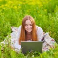 à l'extérieur dans l'herbe se trouve une jeune fille souriante et utilise un ordinateur portable