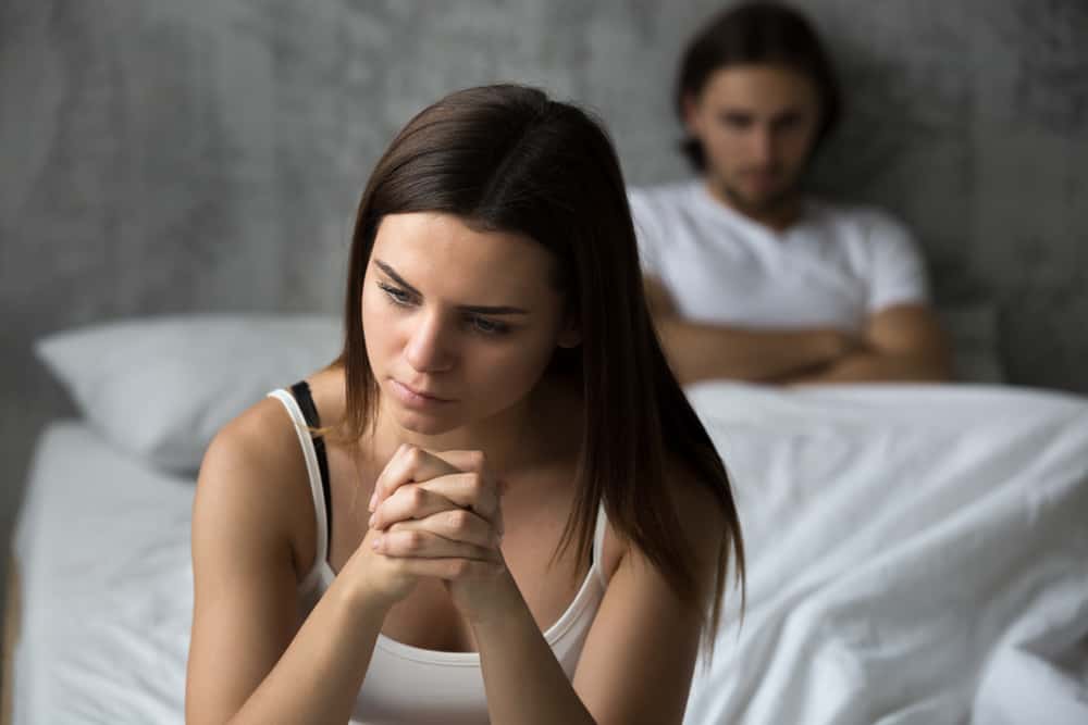 Une femme frustrée assise sur un lit double derrière elle un homme couché