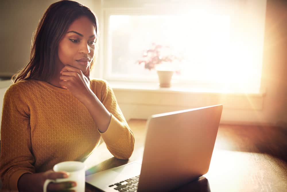 Une femme noire sérieuse dans un T-shirt jaune avec une tasse de café à la main travaille derrière un ordinateur portable