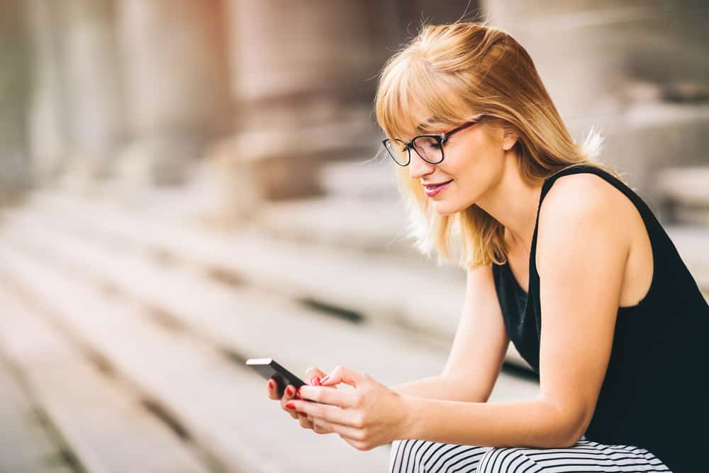 Une femme souriante avec des lunettes se trouve à l'extérieur dans les escaliers et utilise un téléphone portable