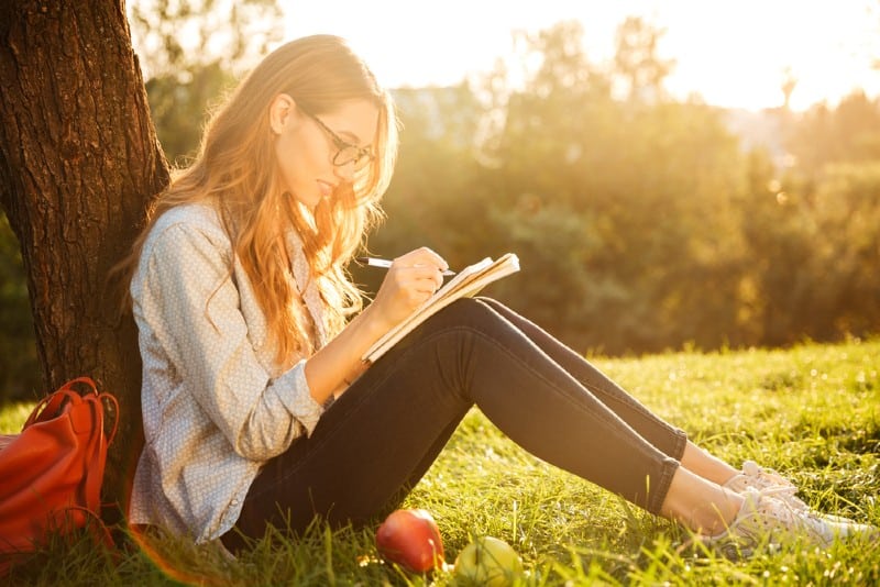 Vue latérale d'une femme brune souriante à lunettes assis près de l'arbre dans le parc et écrire quelque chose sur le cahier