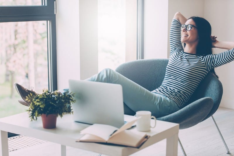 Belle femme gardant les mains derrière la tête et regardant à travers une fenêtre avec le sourire alors qu'il était assis sur son lieu de travail