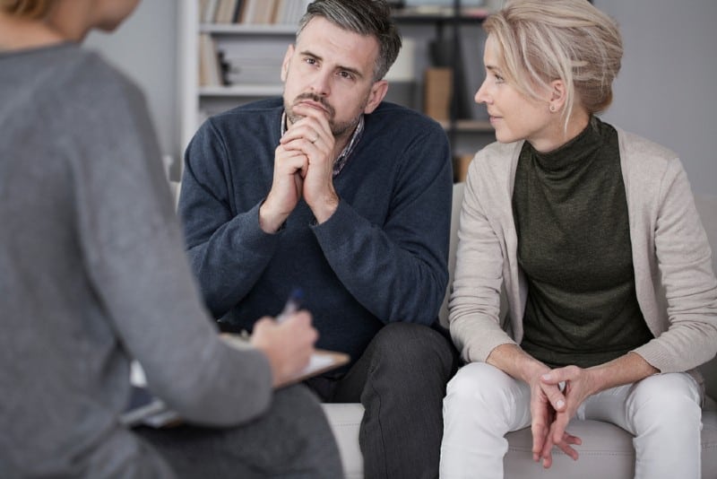 Femme soutenant son mari en thérapie avec l'homme écoutant curieusement le conseiller dans un bureau gris