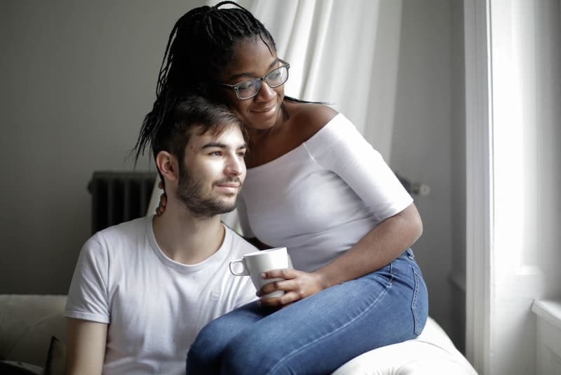 sur le canapé est assise une femme noire sur les genoux de son homme et ils regardent joyeusement par la fenêtre