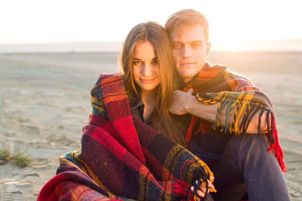 un homme et une femme sont assis près de la plage recouverts d'une couverture