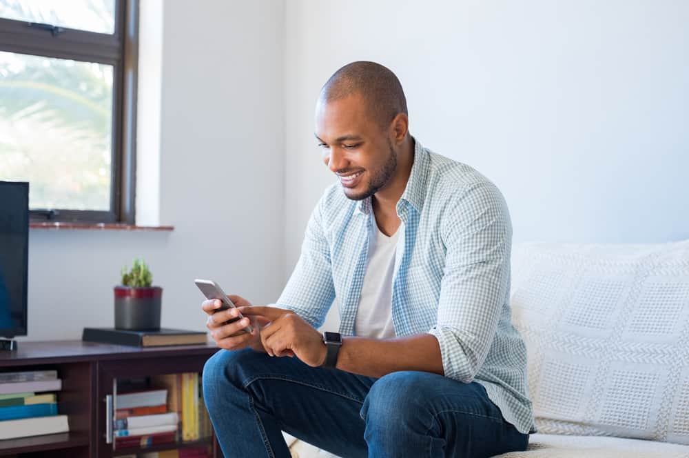 un homme noir souriant est assis sur un canapé et utilise un téléphone portable