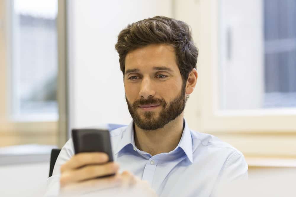un homme souriant avec une barbe est assis et écrit un sms