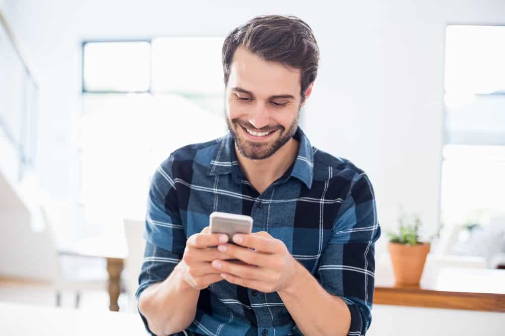 un homme souriant dans une chemise à carreaux écrit un sms dans la maison