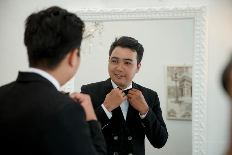 un homme souriant en costume syntonise un nœud papillon devant un miroir