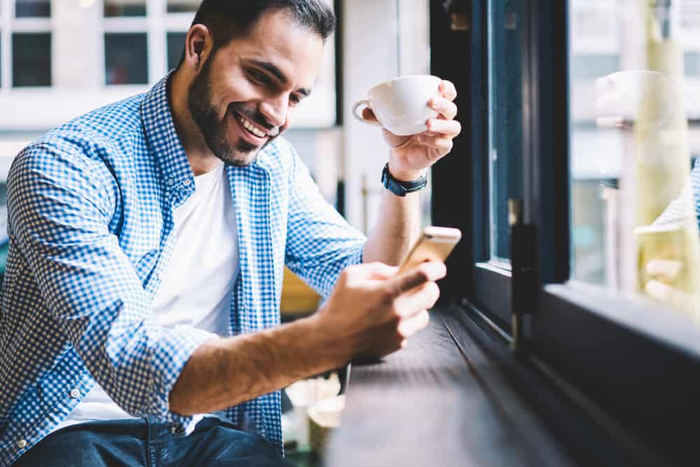 un homme souriant est assis dans un bar près de la fenêtre et utilise un téléphone portable