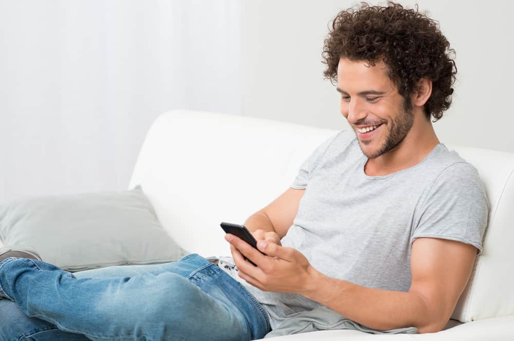 un homme souriant est assis sur le canapé et écrit un sms