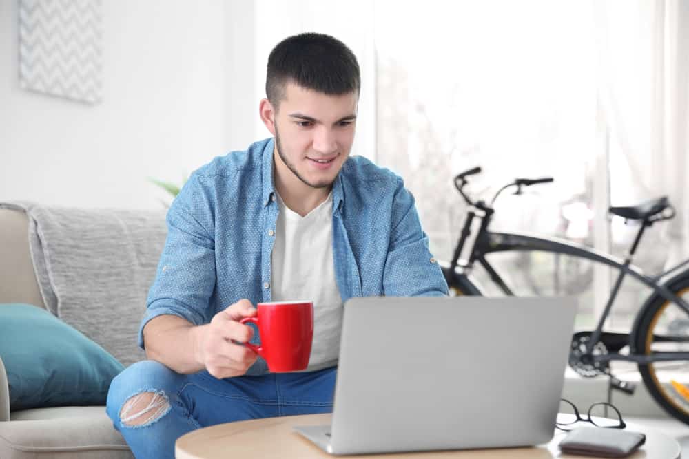 un jeune homme souriant est assis dans la maison et à l'aide d'un ordinateur portable