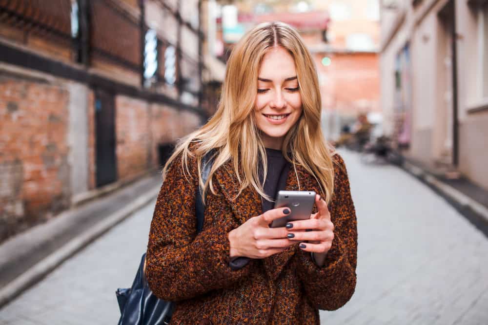 une blonde souriante se tient dans la rue et écrit un sms
