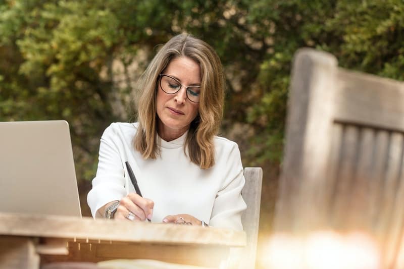 une femme aux cheveux blonds avec des lunettes est assise à une table et écrit