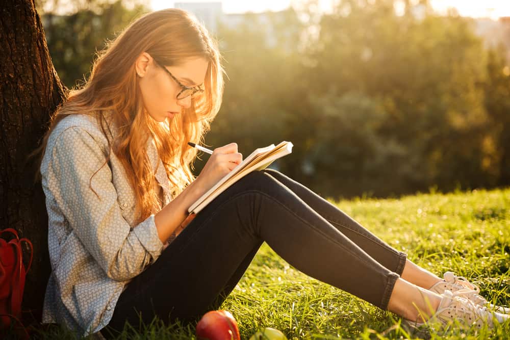 une femme aux cheveux blonds est assise sur l'herbe appuyée contre un arbre et écrit