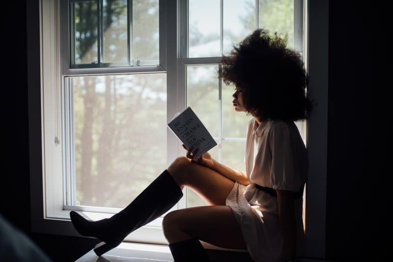 une femme aux cheveux crépus est assise près de la fenêtre et lit un livre