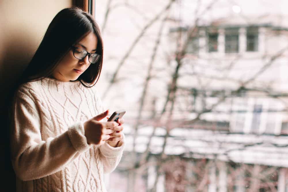 une fille avec des lunettes se tient près de la fenêtre et écrit un sms